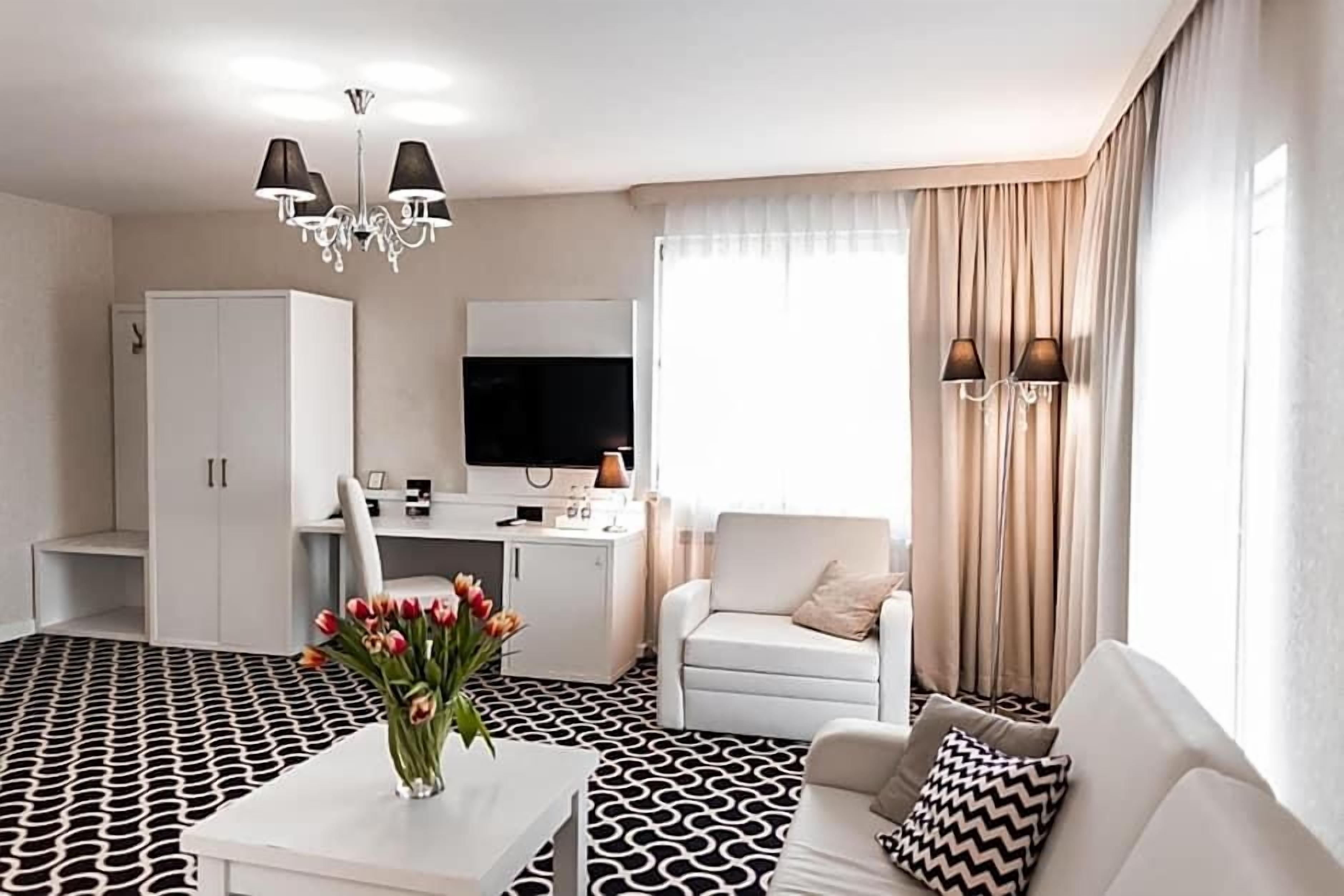 Quarto de hotel moderno com piso em padrão preto e branco, um sofá branco, uma poltrona branca, uma mesa com televisão de tela plana, um guarda-roupa e um lustre.