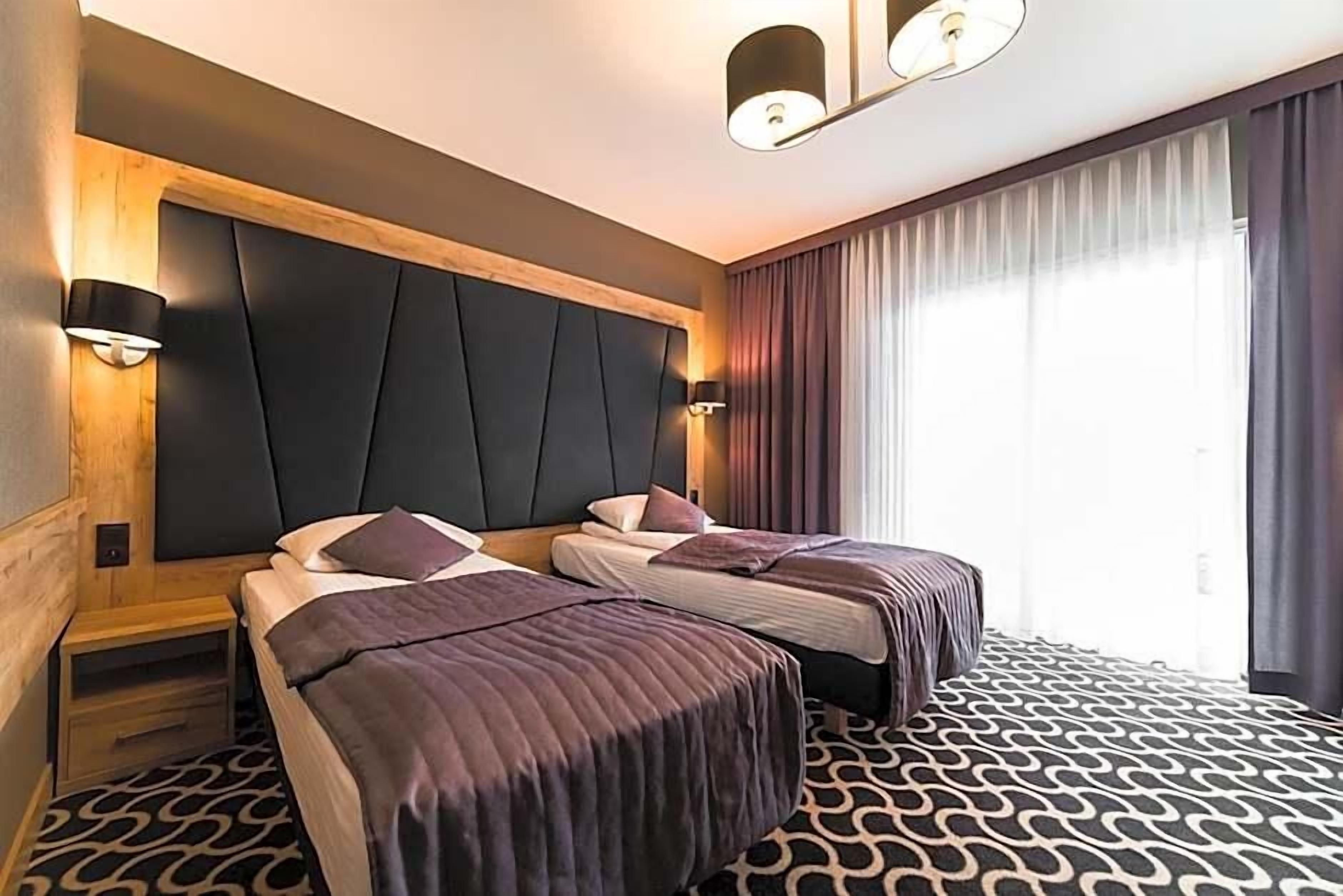 A hotel room with two beds, a large padded headboard, wall-mounted lights, and a patterned carpet.