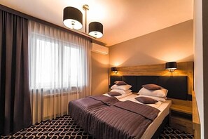 A hotel room with a bed, two bedside lamps, a window with curtains, and a headboard with a light fixture. - Hotel Coloseum (Jaroslaw)