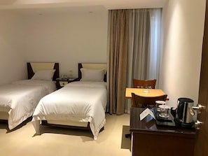 Quarto de hotel com duas camas, uma mesa de jantar, uma cadeira, uma cafeteira e uma mesinha de cabeceira com abajur.