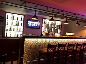 Uma área de bar com balcão, prateleiras cheias de garrafas, luminárias pendentes e uma tela de televisão.
