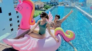A poolside scene with a pink unicorn float, two people, and a cityscape in the background. - Duke Casa Apartment (Da Nang Municipality)