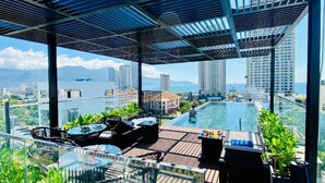 A rooftop terrace with a glass balustrade, outdoor seating, and a view of a cityscape and water body. - Duke Casa Apartment (Da Nang Municipality)