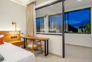 Habitación de hotel con cama, escritorio, silla, mesita de noche, lámpara y un ventanal con vistas al exterior.