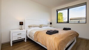 Un dormitorio con cama, mesita de noche blanca, dos lámparas de noche, ventana con vistas y suelo de madera.