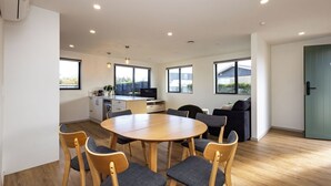 Un interior moderno con una mesa redonda de madera rodeada de sillas, una zona de cocina y ventanas amplias.