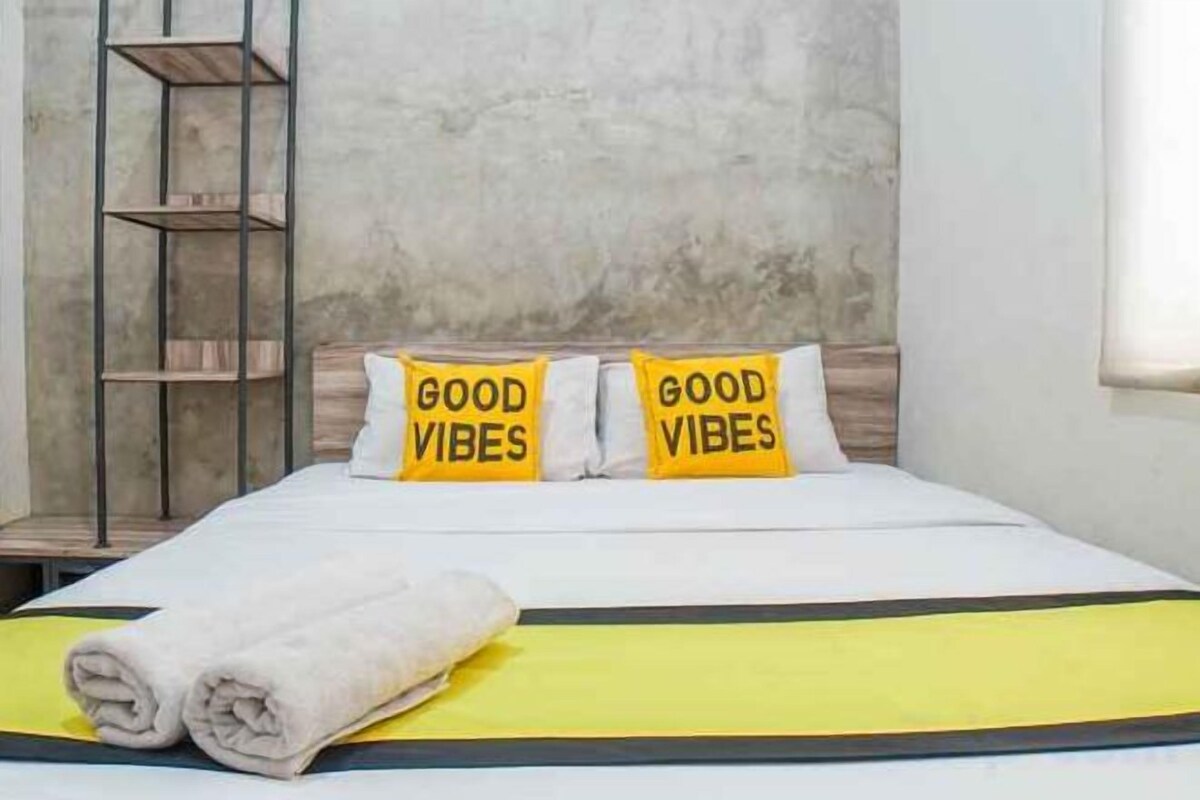 Ein Bett mit Kissen, auf denen 'GOOD VIBES' steht, und einem zusammengerollten Handtuch auf einer gelben Bettdecke.