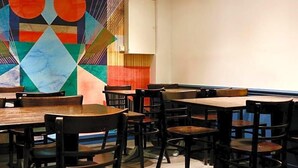 L’intérieur d’un restaurant, avec des tables et des chaises, une fresque murale aux motifs géométriques et un buffet blanc.