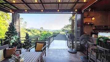 An open-air patio with a view of greenery and hills, furnished with chairs and a table, and a bar area with stools.