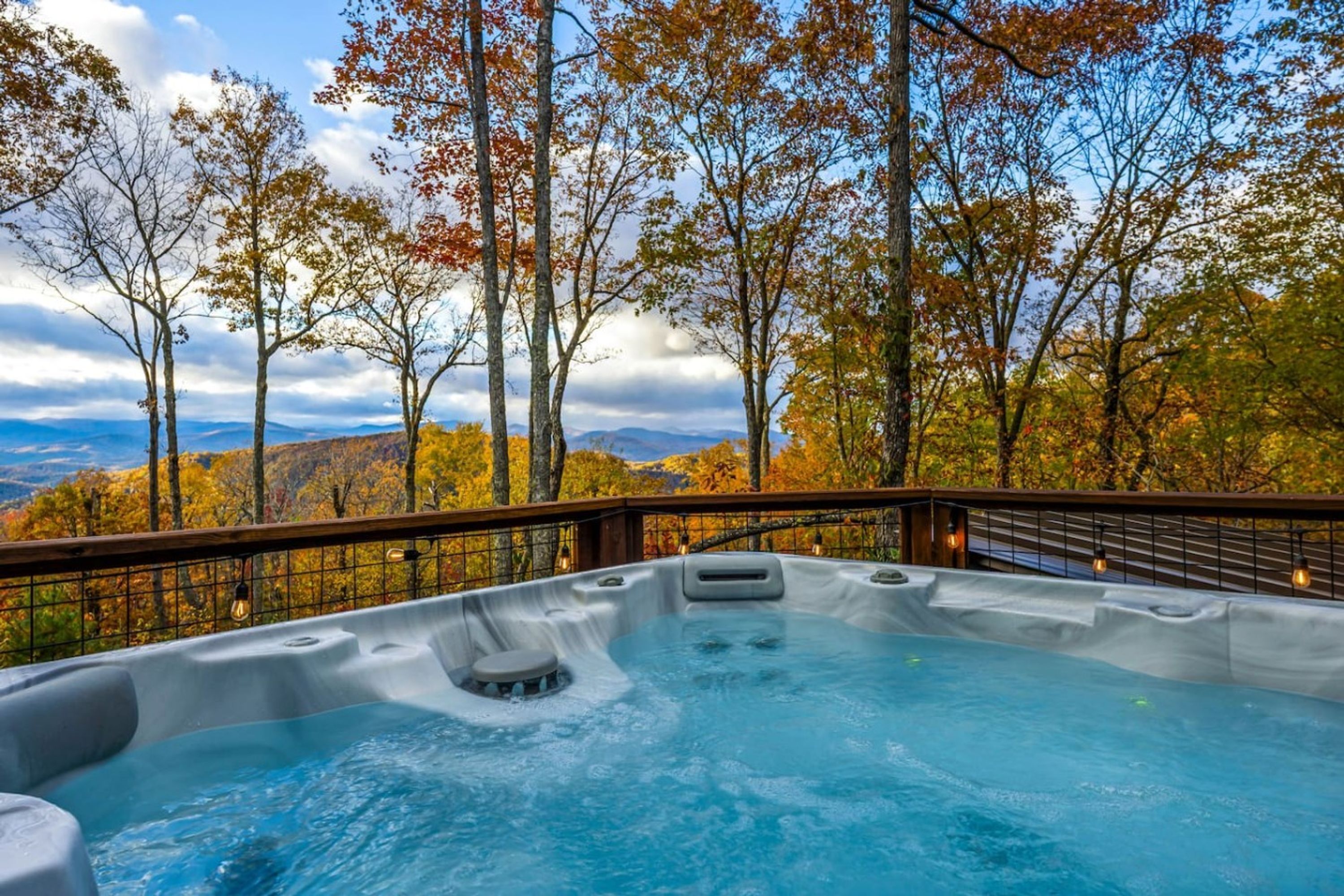 A hot tub with a wooden deck and metal railing, surrounded by trees with autumn foliage and a mountainous backdrop.