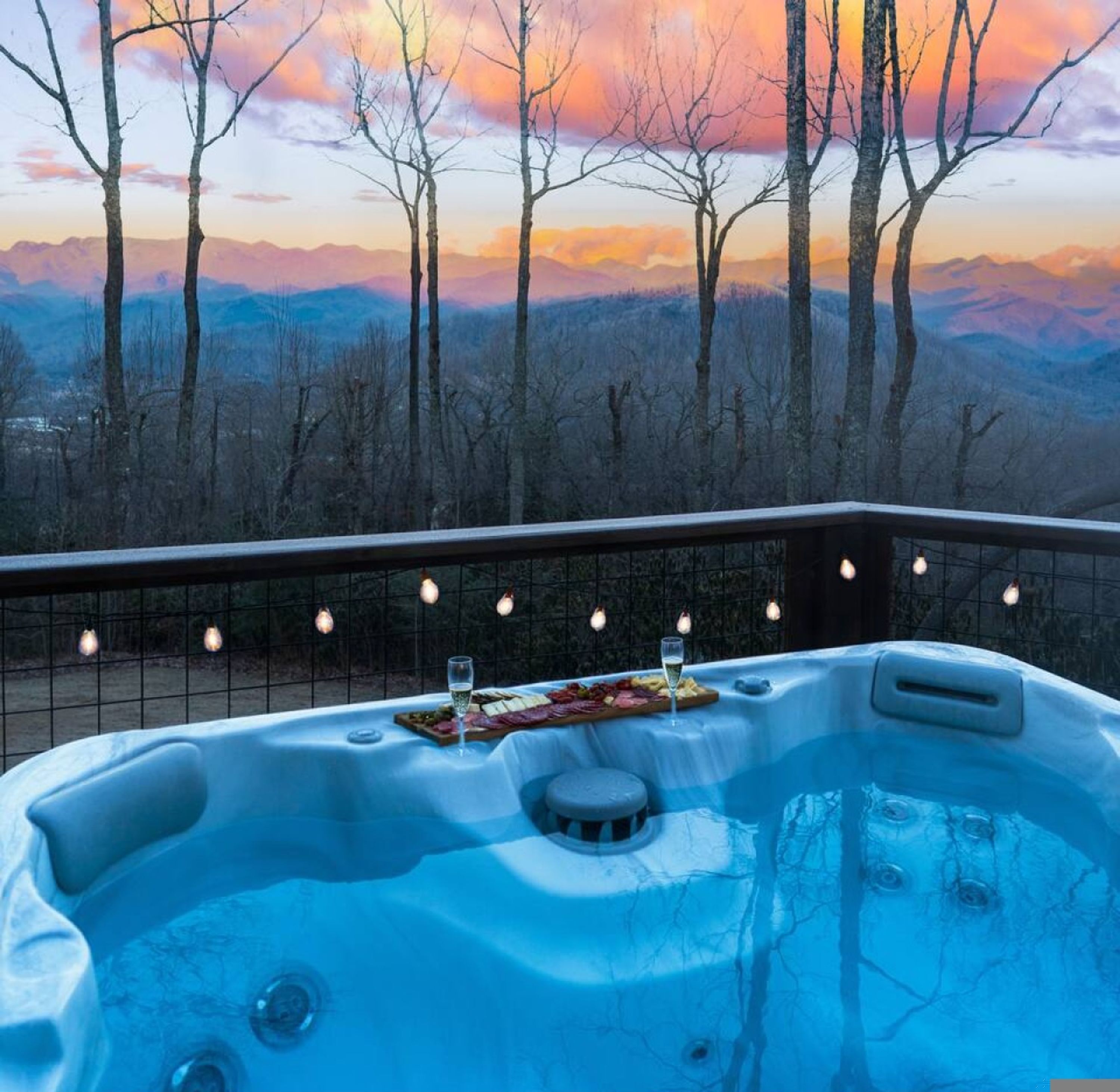 A hot tub with a scenic mountain view, a dining table with food and drinks, and string lights on a balcony.