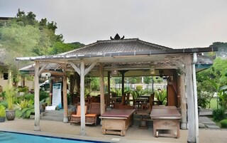 Un espacio al aire libre techado con mesas y sillas, una piscina y un edificio con tejado de azulejos al fondo.