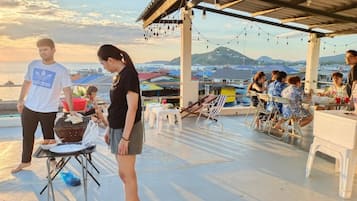 An outdoor dining area with tables and chairs, a grill, and a view of the sea and mountains.