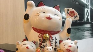 A large cat figurine with smaller cats and bells, set against a backdrop of a bridge and water. - Great House99 (Bangkok)