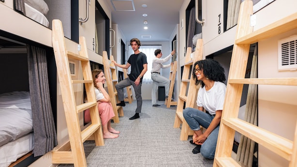 A corridor with bunk beds, a person standing, and another person sitting on the floor. - GRAND HOSTEL LDK Hiroshima (Hiroshima)