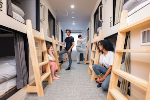 A corridor with bunk beds, a person standing, and another person sitting on the floor.