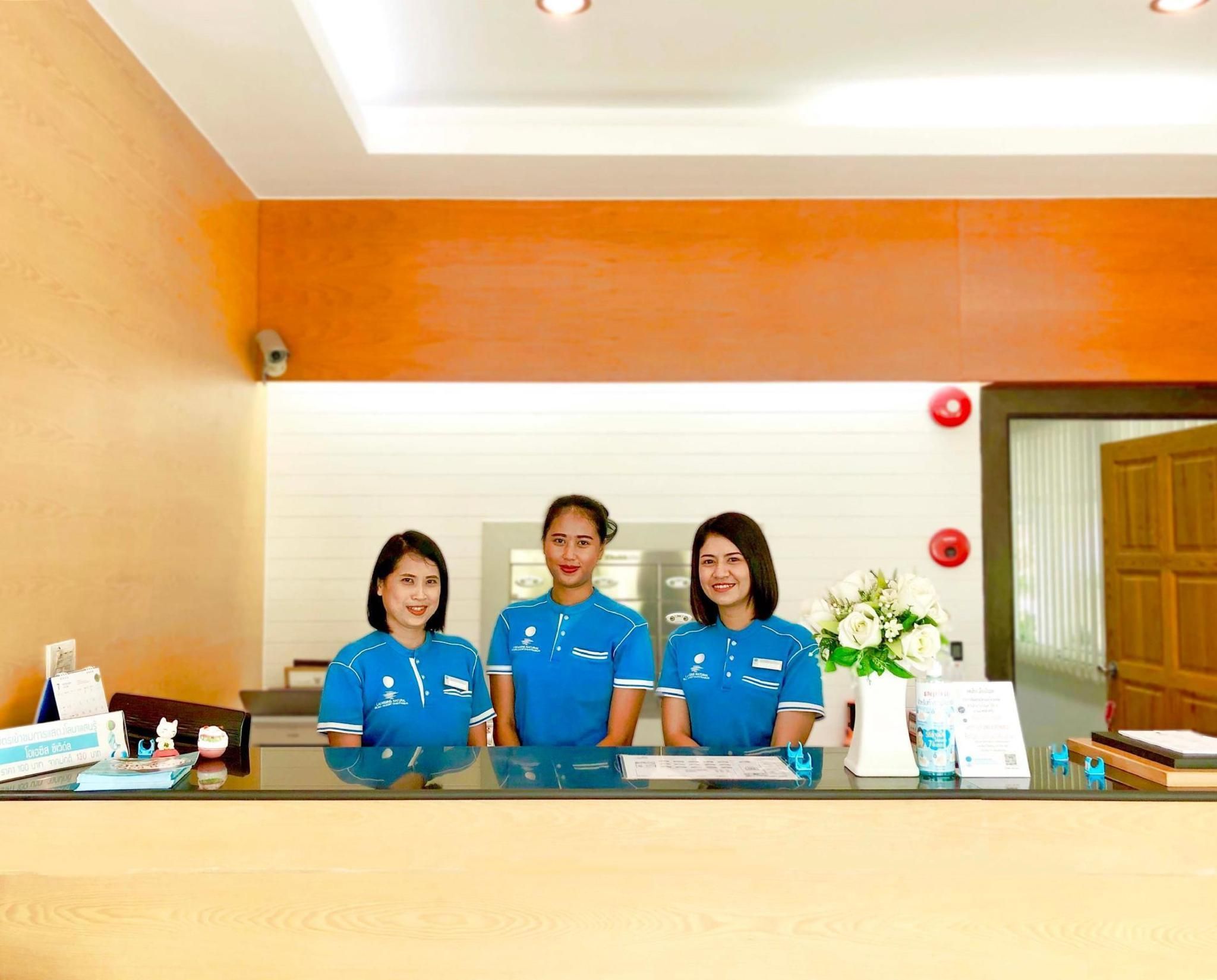 A reception desk with three staff members in blue uniforms, a white flower arrangement, and a wooden door in the background.