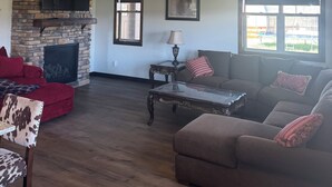 A living room with a stone fireplace, a ceiling fan, a TV mounted on the wall, and a window with a view of the outdoors. - Kara Creek Ranch - 2 Bed/2 Bath - Cabin 11 (Sundance)