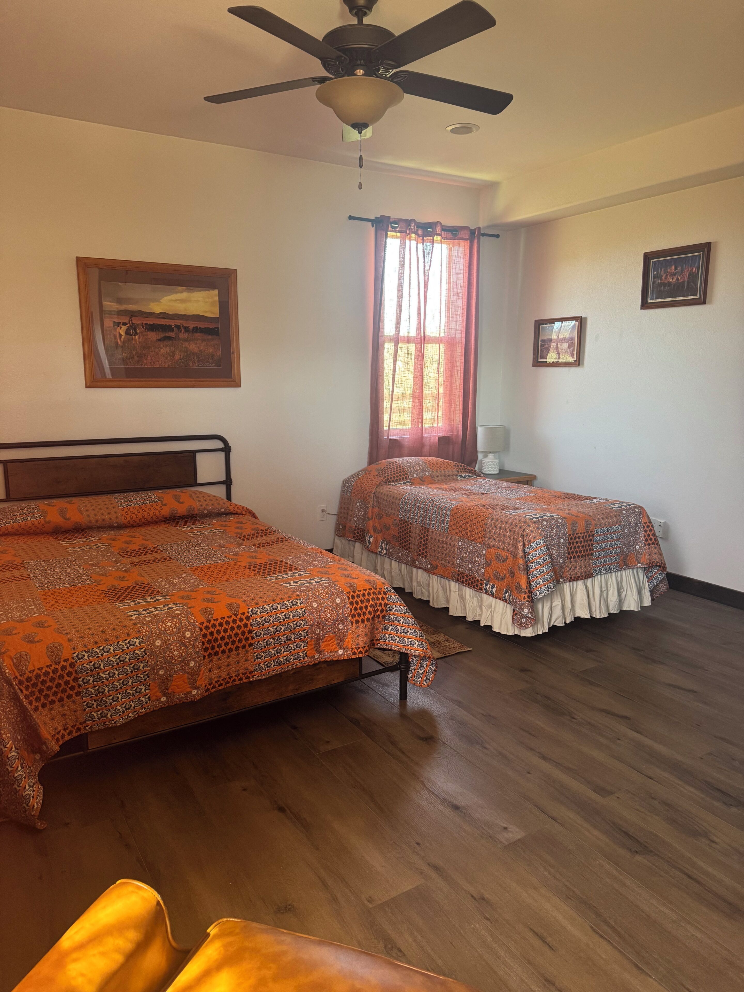 Una habitación con dos camas, un ventilador de techo, una ventana con cortinas y un cuadro en la pared.