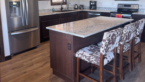 A modern kitchen with dark wooden cabinets, stainless steel appliances, a granite island, and bar stools. - Kara Creek Ranch - 2 Bed/2 Bath - Cabin 11 (Sundance)