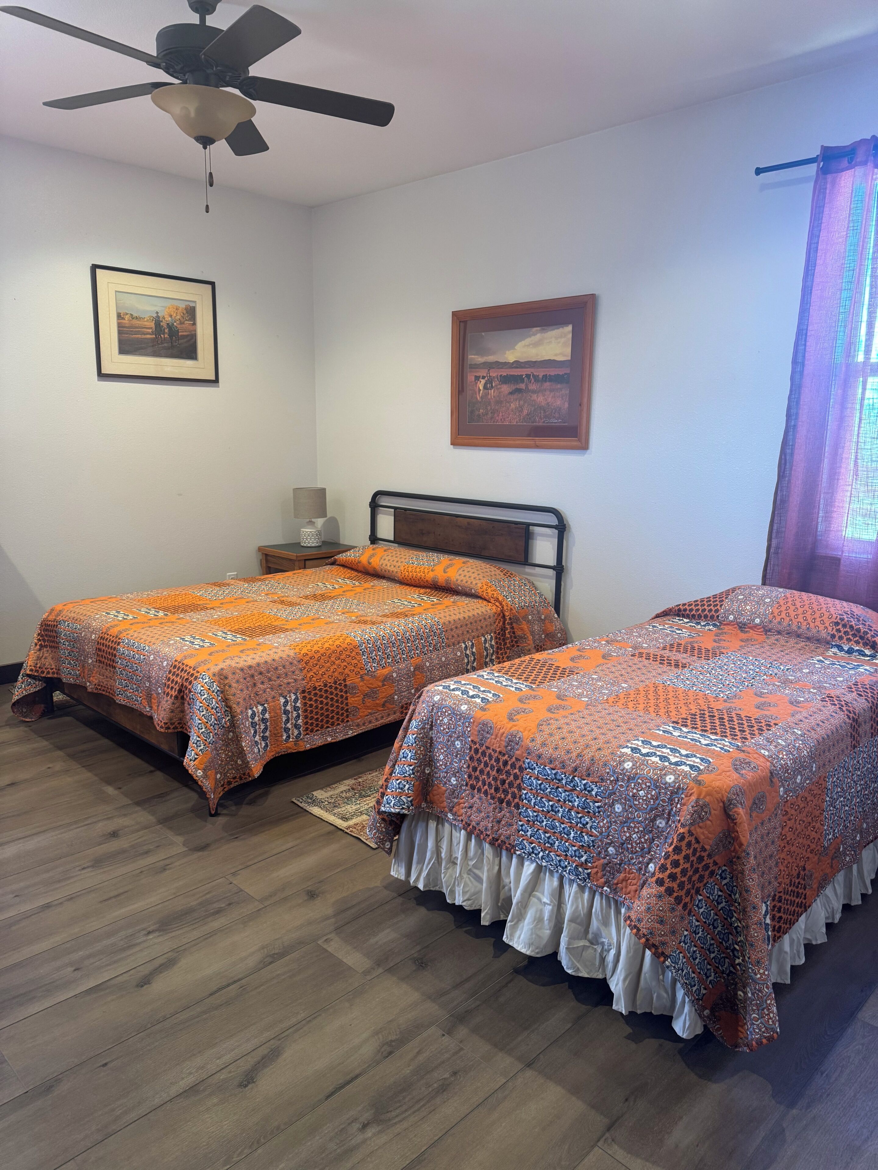 Un dormitorio con dos camas, un ventilador de techo, cuadros en la pared y una ventana con cortinas.