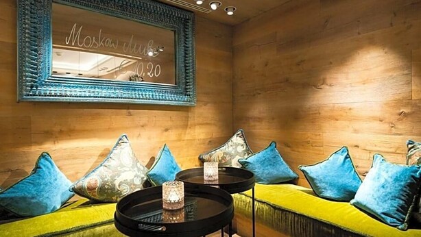 Ein gemütliches Zimmer mit Holzwänden, einem gelben Sofa mit blauen Kissen, zwei kleinen runden Tischen und einem Spiegel mit Schriftzug.