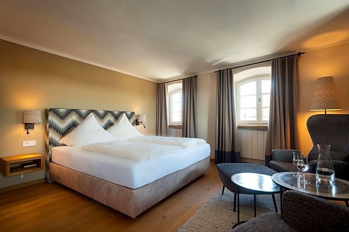 A hotel room with a large bed, a small table with glasses, a chair, and a lamp.