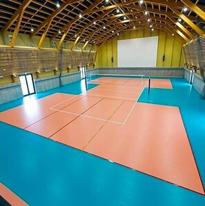 Una cancha deportiva interior con piso azul y marcas naranjas, rodeada por una estructura de madera con techo alto.