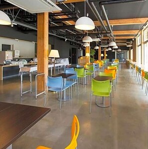 Una cafetería amplia con techos altos, vigas a la vista y una variedad de sillas y mesas de colores.