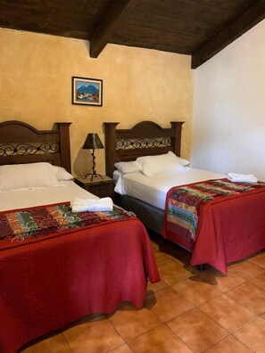 A hotel room with two beds, each with a red bedspread, a nightstand with a lamp, and a painting on the wall. - Hotel T Kon T (Antigua Guatemala)