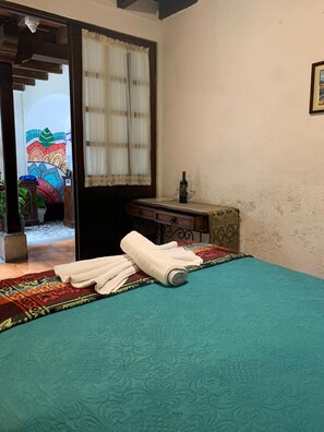 A room with a bed, a small table with a bottle and glasses, a window with curtains, and a colorful door. - Hotel T Kon T (Antigua Guatemala)