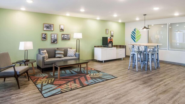 A living room with a sofa, armchair, coffee table, and a patterned rug. There is a dining area with a bar stool and a small table. The room has a green wall and ceiling lights. - Woodspring Suites Jacksonville Campfield Commons (Jacksonville)