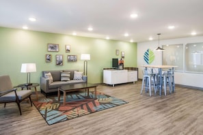 Un salon comprenant un canapé, un fauteuil, une table basse et un tapis à motifs. On y trouve également un coin repas avec un tabouret de bar et une petite table. La pièce est dotée d’un mur vert et d’un éclairage au plafond.