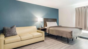 A hotel room with a bed, a sofa, a nightstand, a lamp, and a window with curtains. - Woodspring Suites Jacksonville Campfield Commons (Jacksonville)