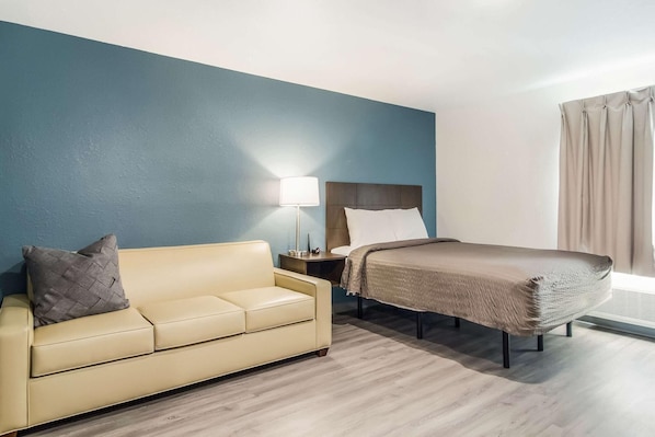 Habitación de hotel con cama, sofá, mesita de noche, lámpara y ventana con cortinas.