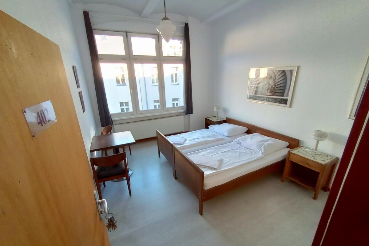 Une chambre d’hôtel avec un lit double, un bureau, une chaise, une table de chevet, une lampe et une fenêtre avec des rideaux.