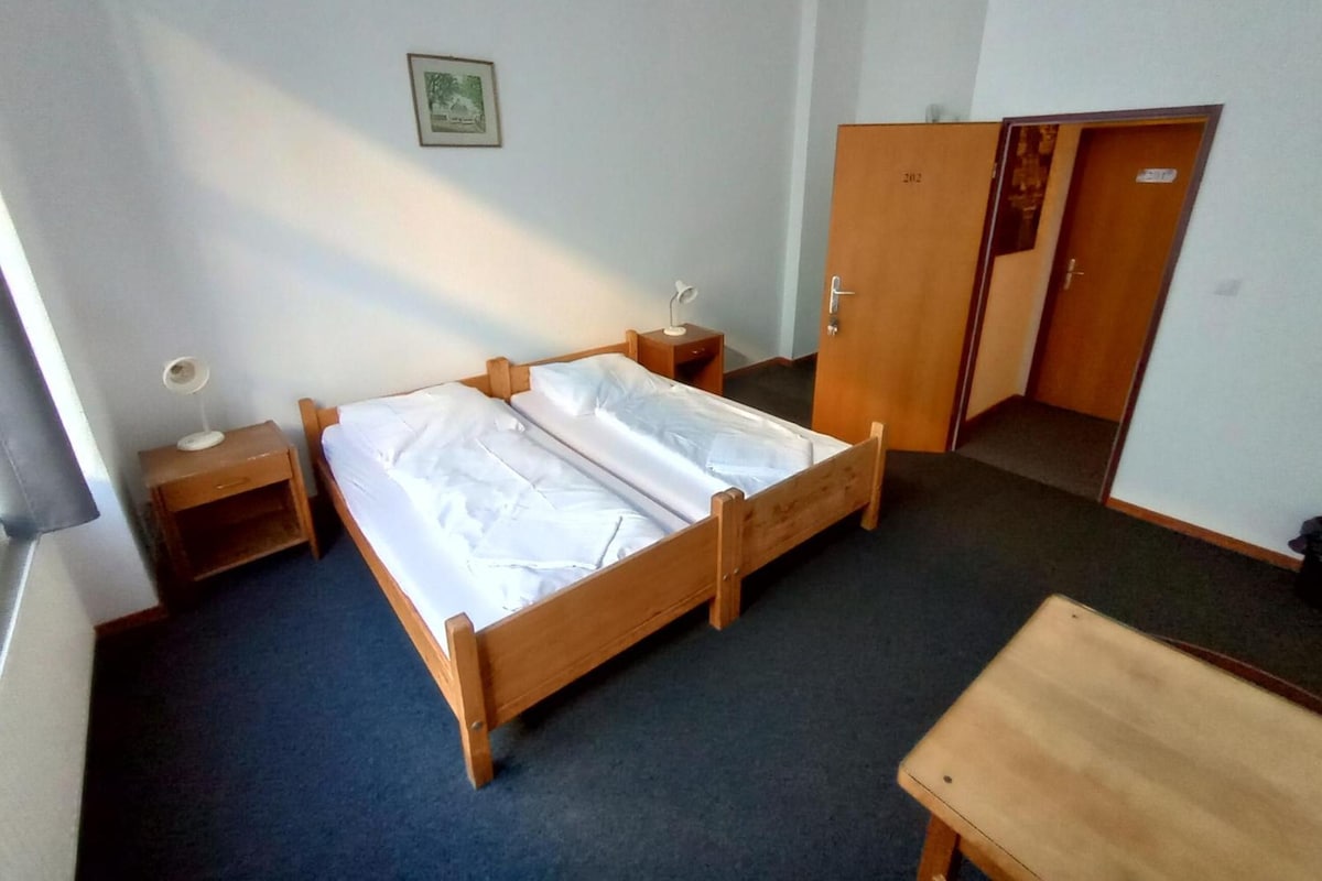 Ein Zimmer mit einem Einzelbett, einem hölzernen Bettgestell, einem Nachttisch mit Lampe, einem Kleiderschrank und einem kleinen Schreibtisch mit Stuhl.