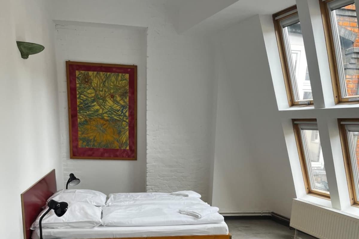 Ein Zimmer mit einem Bett, einem kleinen Tisch, einer Lampe, einem Bild an der Wand und einem Fenster.
