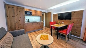 Un espace de vie compact comprenant un coin cuisine, un coin repas avec des chaises rouges et un téléviseur fixé au mur.