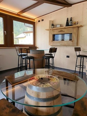 A dining area with a glass-top table, wooden barrel centerpiece, and bar stools. - Soggiorno Dolomiti (Mazzin)