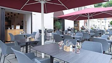 Ein Straßencafé mit Tischen, Stühlen, Sonnenschirmen und einer Theke mit Auslagen.