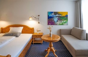 A hotel room with a bed, a sofa, a small table with a vase, a nightstand, a lamp, and a wall with a colorful painting. - Hotel Goldener Hirsch (Kaufbeuren)