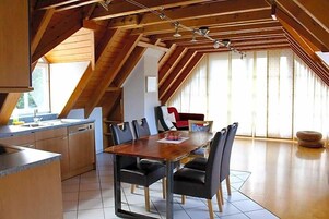 A dining area with a wooden table and chairs, a kitchen with a dishwasher, and a large window with curtains.