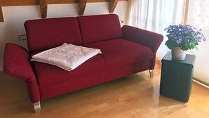 Ein rotes Sofa mit einem weißen Kissen in einem Raum, der Holzbalken, einen Blumentopf und ein Fenster mit Vorhängen zeigt.