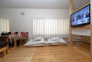 A room with a wooden floor, a television mounted on the wall, a bed with white bedding, and a table with flags.