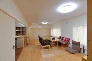 A spacious living area with a wooden floor, a sofa set, a dining table, and a kitchen area in the background.