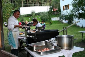 Uma área de cozinha ao ar livre com churrasqueira, um chef e uma mesa com comida.