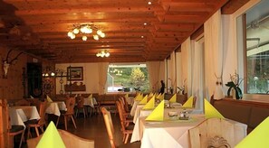 Um restaurante com vigas de madeira no teto, mesas com toalhas amarelas e vista para o verde lá fora.