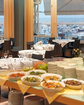 Een restaurant met meerdere tafels gedekt voor diners, een buffet met diverse gerechten, en uitzicht op de jachthaven.
