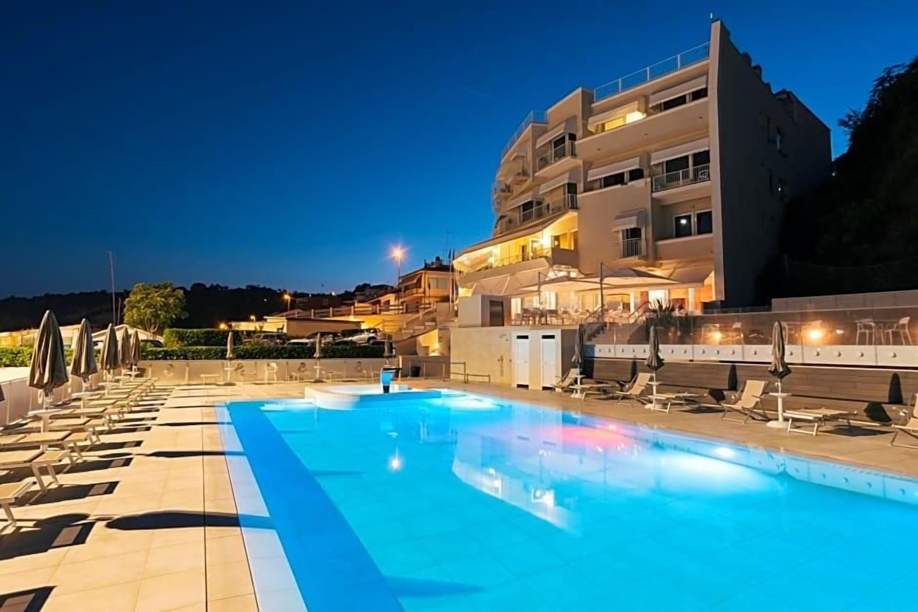 A swimming pool with lounge chairs and umbrellas, a multi-story building with balconies, and a clear sky.